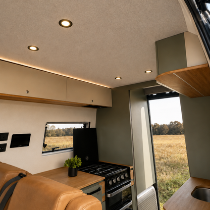 The interior of a modern motorhome or camper van, featuring a compact kitchenette with a black gas stove and oven, light wood countertops, a tan leather seat in the foreground, and an open window looking out onto a grassy field.
